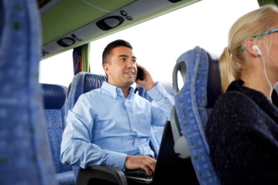 Un homme d'affaires souriant travaille sur son ordinateur portable et passe un appel téléphonique à bord d'un autocar confortable, illustrant les avantages pratiques et économiques de la location bus avec chauffeur pour le transport de groupe.