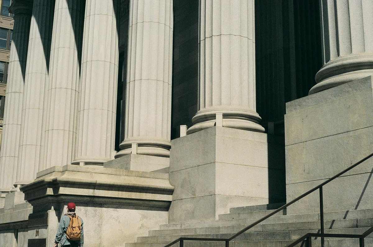 Colonnades blanches d'un édifice, marches en pierre, individu avec sac à dos